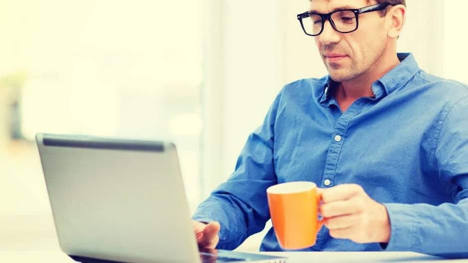 Man in blue shirt in front of laptop drinking coffee